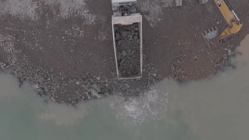 Aerial top-down view of a dump truck unloading large rocks and soil into the water to reinforce a shoreline at a construction site