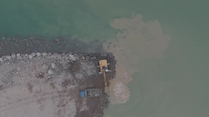 Aerial top-down view of an excavator on a rocky pier, digging and stirring up sediment in the water
