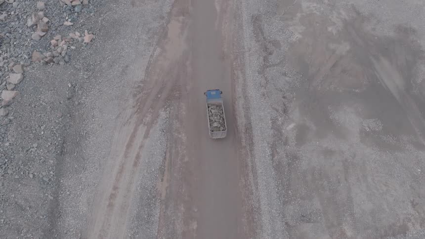 An overhead aerial view follows a large dump truck as it slowly transports a heavy load of rocks along a dusty construction road