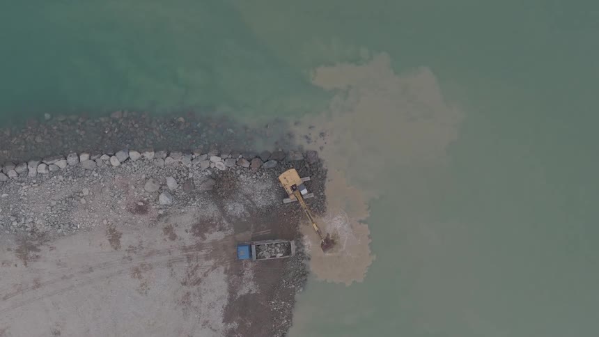 Top-down aerial view of an excavator digging sediment from the water, creating a muddy plume