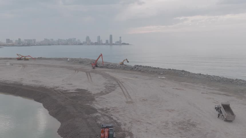 A wide aerial shot captures a massive coastal construction project with excavators and trucks shaping a new peninsula against a distant city skyline