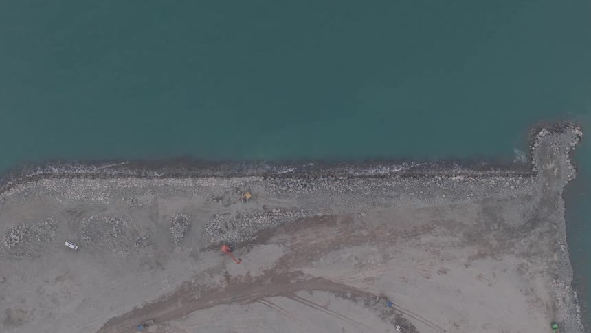 An overhead drone shot captures the calm waves of the turquoise sea meeting the rocky breakwater of a coastal construction project