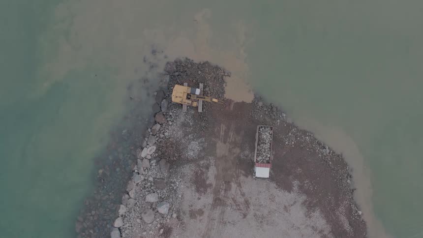 An aerial top-down view shows an excavator moving rocks from a dirt peninsula to build a breakwater into the murky water