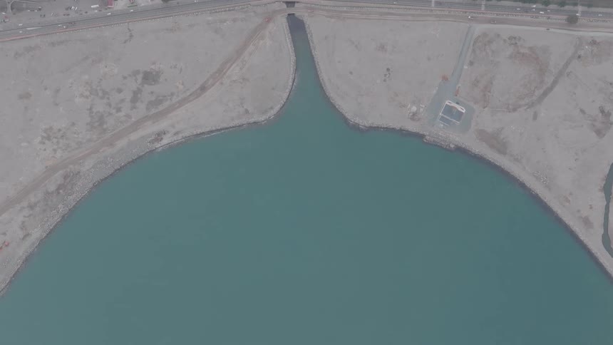 Top-down aerial footage slowly zooms into a man-made peninsula and inlet, with a river flowing under a coastal highway into the sea