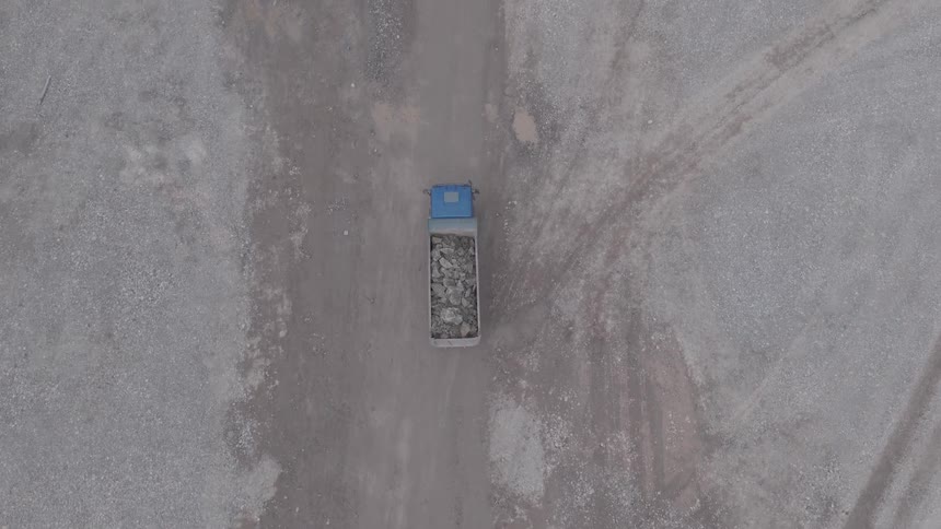 Aerial top-down view follows a loaded dump truck driving through a dusty industrial quarry site