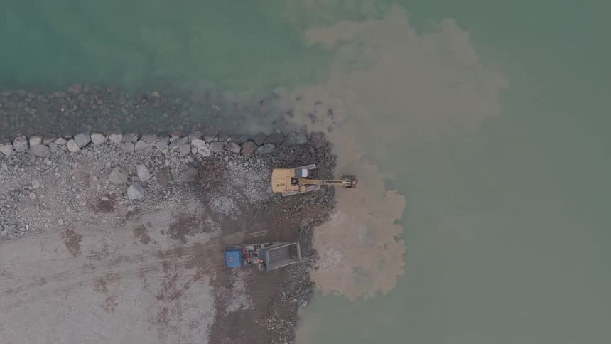 Top-down aerial view of a yellow excavator dredging material from the water and creating a splash on a rocky pier