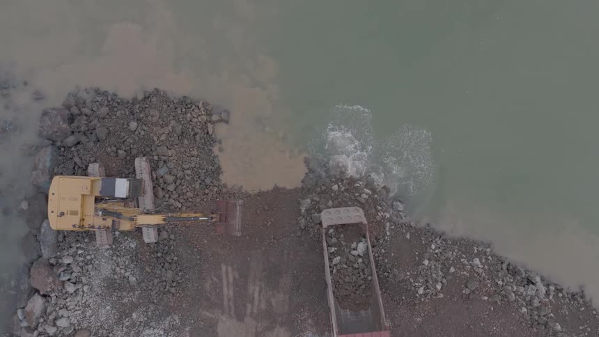 Aerial top-down view of an excavator dumping rocks from a truck into the water, working on a shoreline construction project