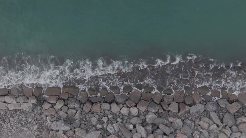 An aerial top-down view captures tranquil ocean waves washing over a rocky shore before the camera slowly ascends