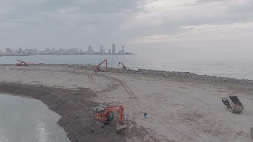 Aerial view of a large-scale coastal construction site with excavators and heavy machinery working on land reclamation with a city skyline in the background