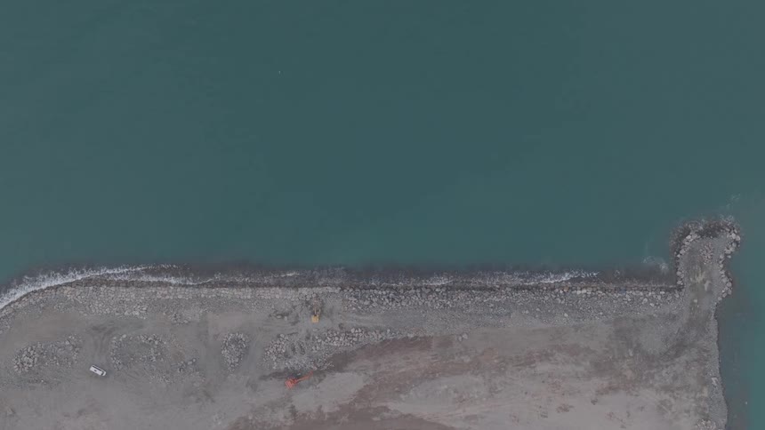 Aerial top-down shot of a coastal construction site where a bulldozer fortifies a new rock breakwater against the calm teal ocean
