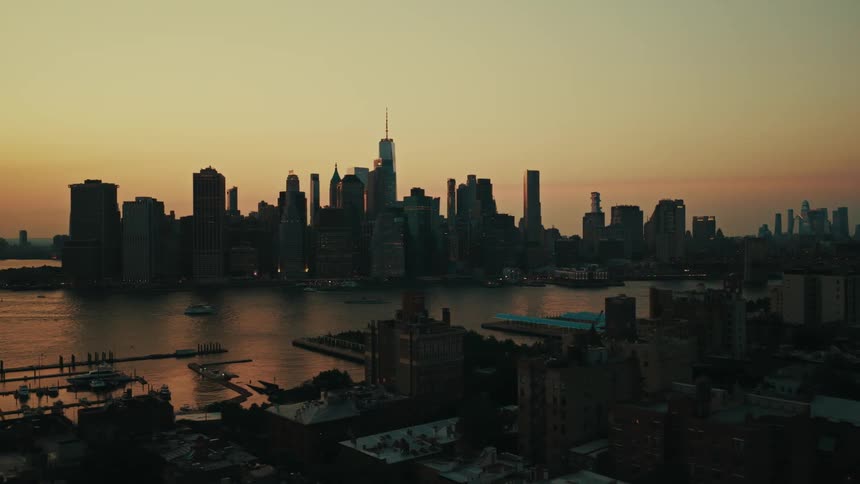 An aerial view reveals the iconic Lower Manhattan skyline silhouetted against a beautiful orange sunset sky over the East River