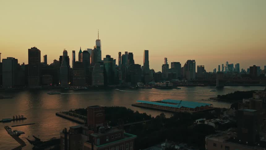 An aerial view pulls back from the iconic Lower Manhattan skyline across the East River at sunset