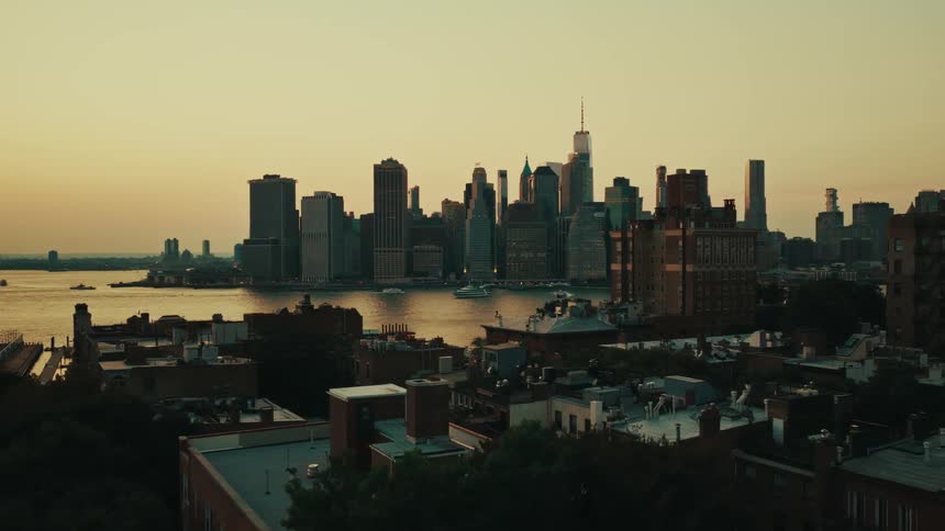 An aerial view pulls back over Brooklyn rooftops to reveal the iconic Manhattan skyline across the East River during a beautiful golden hour sunset