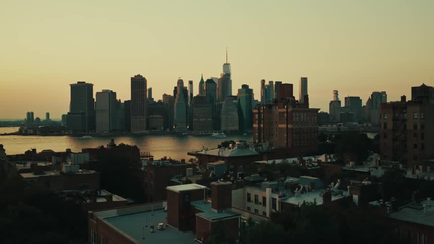 An aerial view pushes forward over the rooftops of Brooklyn towards the Manhattan skyline across the East River at sunrise