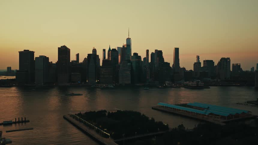 An aerial view pulling back from the silhouetted Lower Manhattan skyline over the East River at sunset, revealing the Brooklyn waterfront