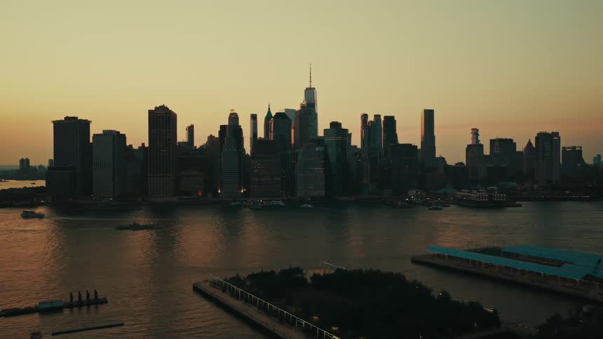 An aerial drone shot slowly pushes forward over the East River towards the silhouetted Manhattan skyline during a beautiful sunset