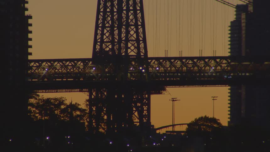A silhouetted train travels across a large steel truss bridge at sunset, framed by city buildings