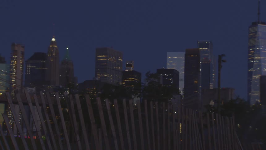 The Lower Manhattan skyline glows at dusk with the iconic One World Trade Center tower visible behind a wooden slat fence