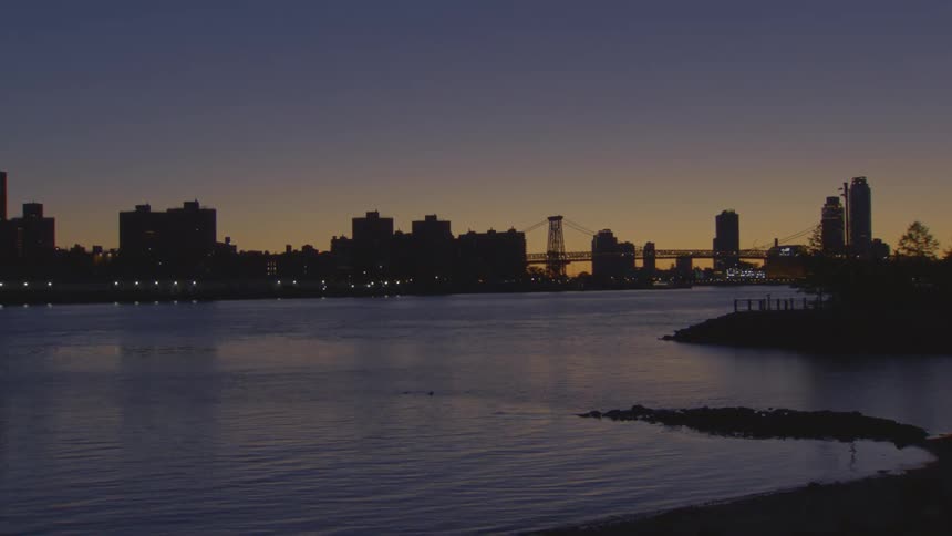 A serene, wide shot of the New York City skyline and Williamsburg Bridge silhouetted against the sunset over the East River at dusk