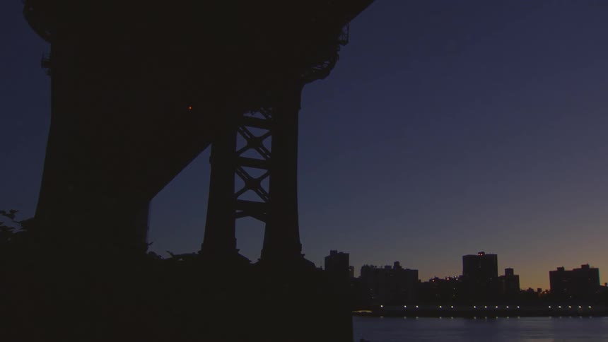 A subway train crosses a bridge into the city skyline at dusk, seen from beneath another bridge's silhouette