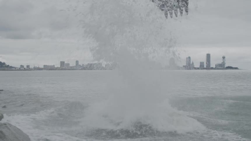 In slow motion, an excavator bucket drops a massive rock into the ocean, creating a huge splash with a city skyline in the background