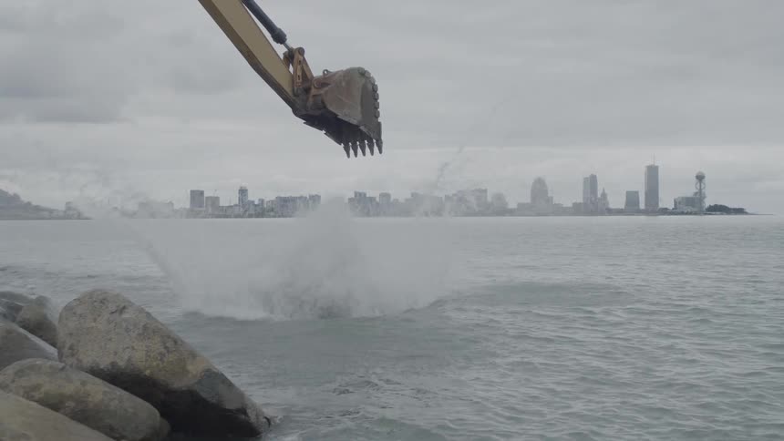An excavator drops a large boulder into the sea in slow motion, creating a big splash with a city skyline in the background