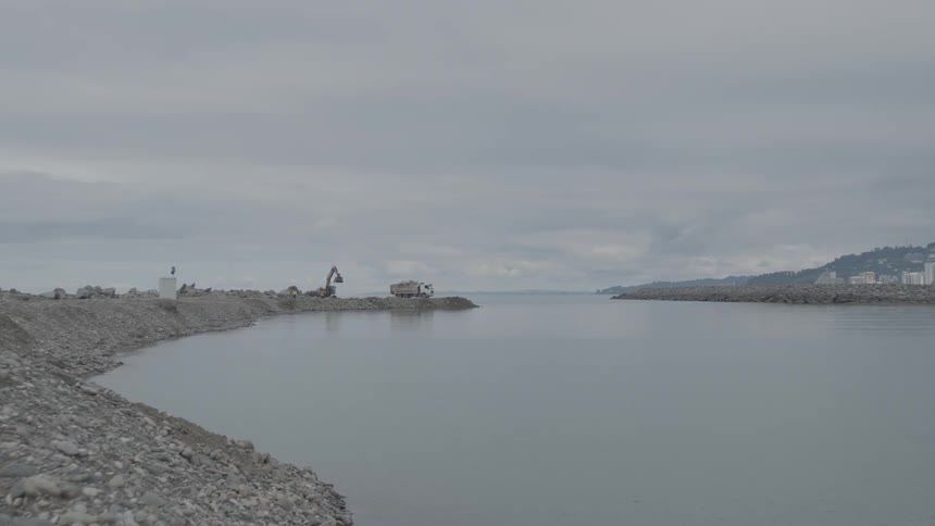 Construction work with an excavator and a truck on a coastal breakwater under a gray, cloudy sky