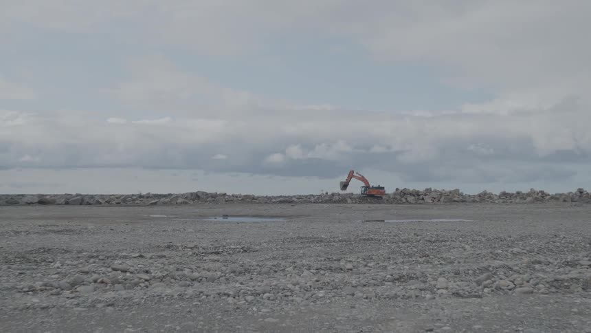 A lone orange excavator works on a rock wall in a vast gravel lot under a cloudy, grey sky