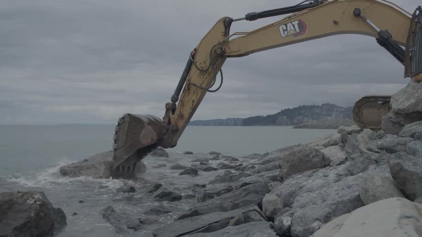 A yellow excavator carefully places a large rock into the ocean to reinforce the coastline on an overcast day