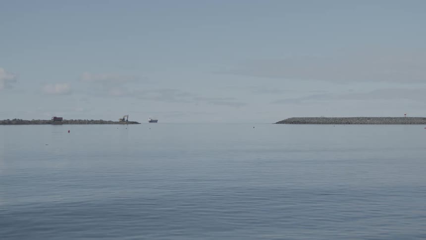 A distant excavator diligently constructs a rocky breakwater in the vast, tranquil blue sea