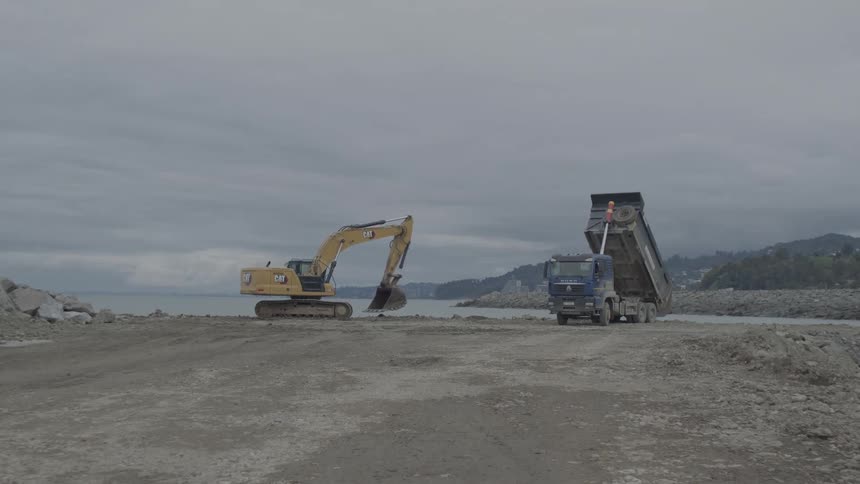 A dump truck unloads dirt at a coastal construction site next to a stationary excavator under an overcast sky