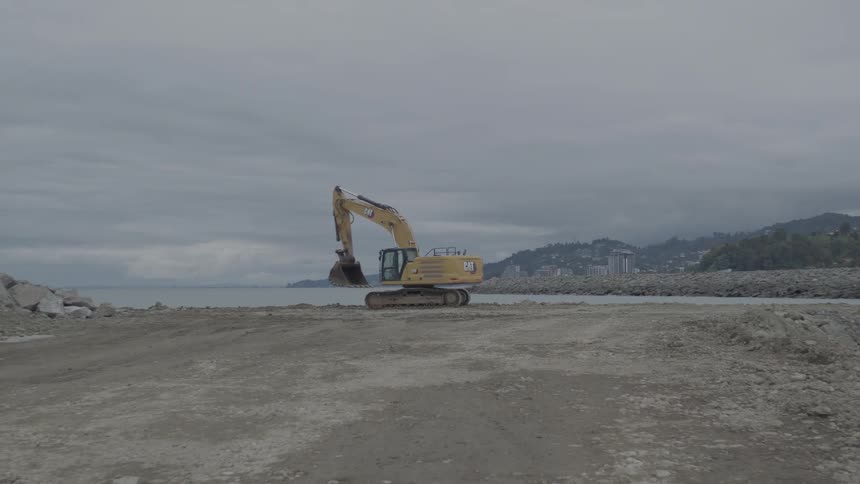 A yellow excavator operates on a desolate coastal construction site next to the sea on an overcast day