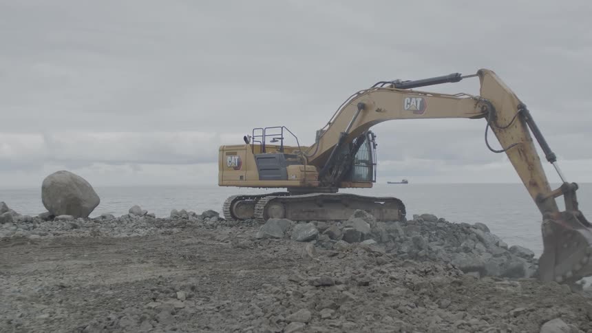 A yellow excavator works on a rocky shoreline, moving large stones to reinforce the coast on a cloudy day