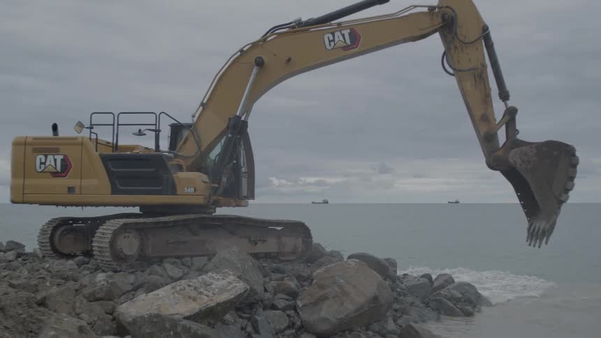 A large yellow excavator meticulously moves and places large boulders into the sea to construct a breakwater on a cloudy day