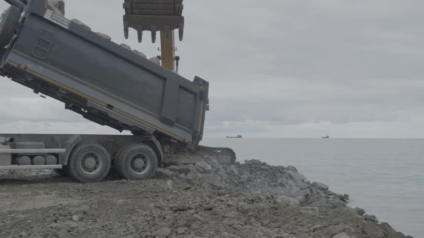 An excavator and a dump truck work in tandem to unload large rocks onto a breakwater in slow motion
