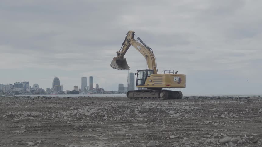A yellow excavator slowly moves into position on a rocky construction site with a modern city skyline across the sea in the background