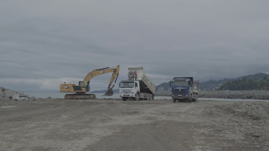 A wide static shot shows an excavator loading a dump truck while another unloads its cargo at a seaside construction site under a cloudy sky