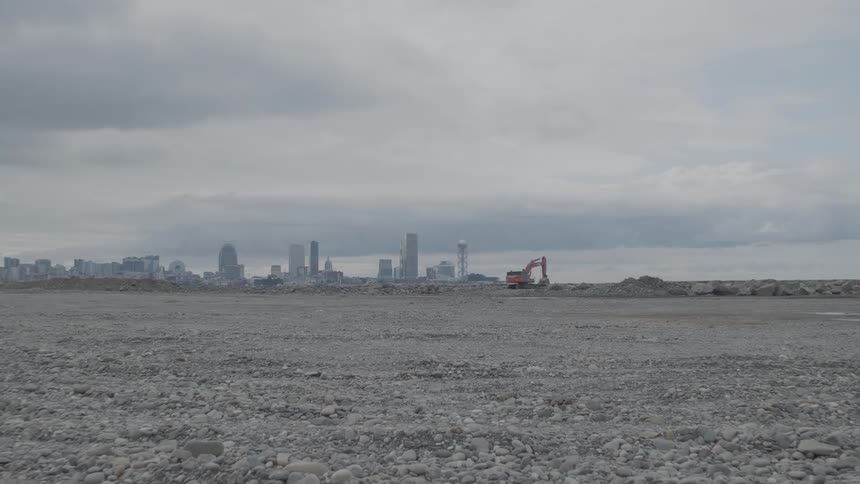 An excavator works on a vast, rocky construction site with a distant city skyline under a heavy, overcast sky