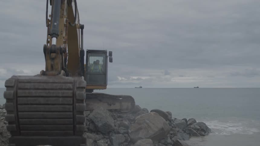A yellow excavator skillfully moves large boulders to construct a breakwater on the ocean shore under a cloudy sky