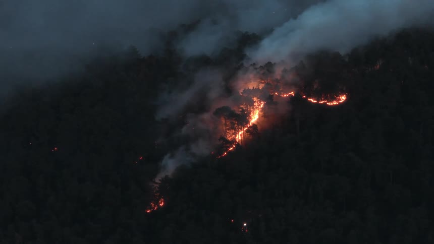 An intense forest fire burns uncontrollably along a dark mountainside at night, sending thick smoke into the foggy air