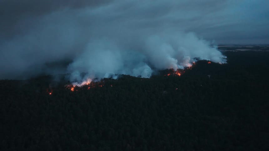 Aerial view of a vast forest fire raging at dusk, with thick smoke rising against the dark, cloudy sky