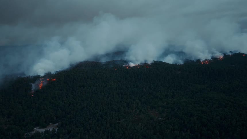 Aerial view of a devastating forest fire raging on a mountainside at dusk, with thick smoke filling the sky