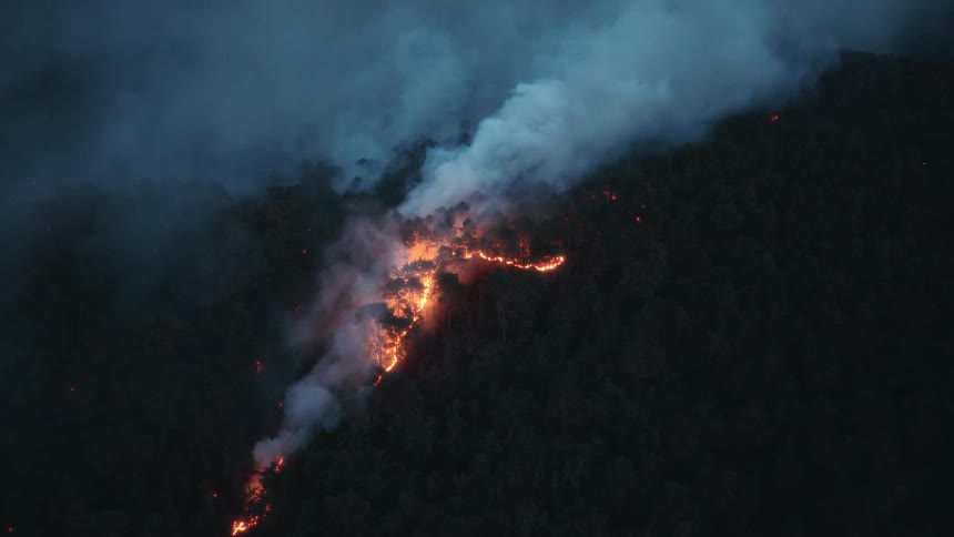 An ominous aerial view shows a wildfire burning a fiery path through a dense forest on a mountainside at night