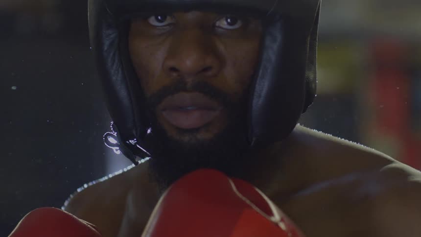 An intense male boxer with a beard stares directly at the camera in a dramatically lit, slow-motion close-up