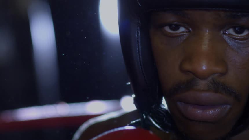A dramatic slow-motion close-up of a focused boxer in headgear staring into the camera as a lens flare engulfs the shot