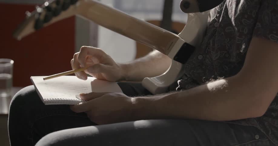 A man taps a pencil on a book while holding a guitar
