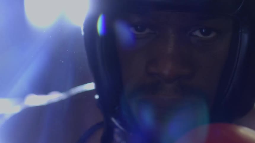 An intense close-up of a focused male boxer in headgear staring directly into the camera with dramatic backlighting
