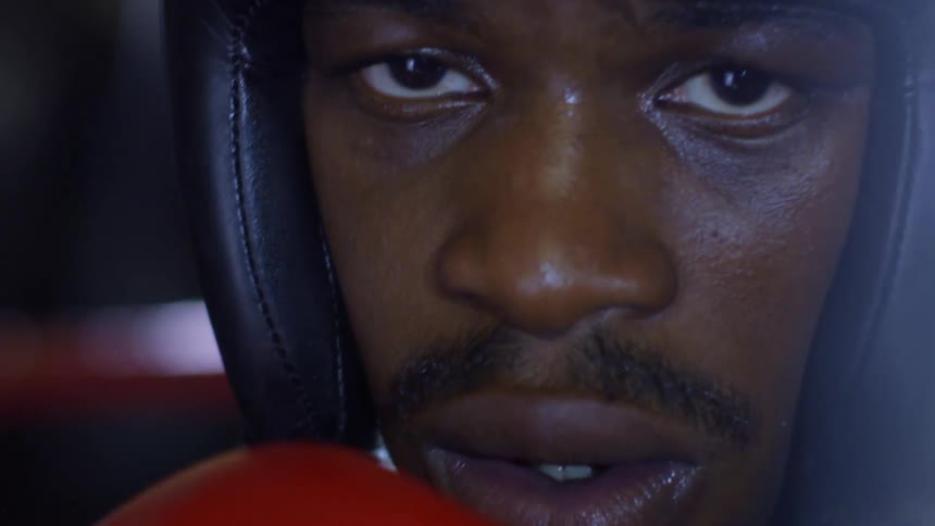 An intense close-up reveals a determined boxer with headgear on, staring forward with dramatic lighting