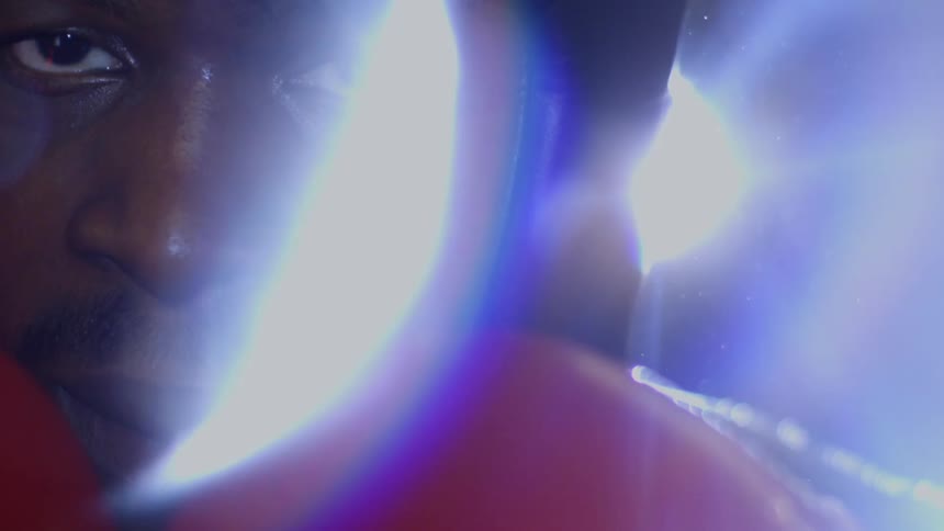 A dramatic close-up in slow motion reveals the intense stare of a determined boxer in red gloves and black headgear