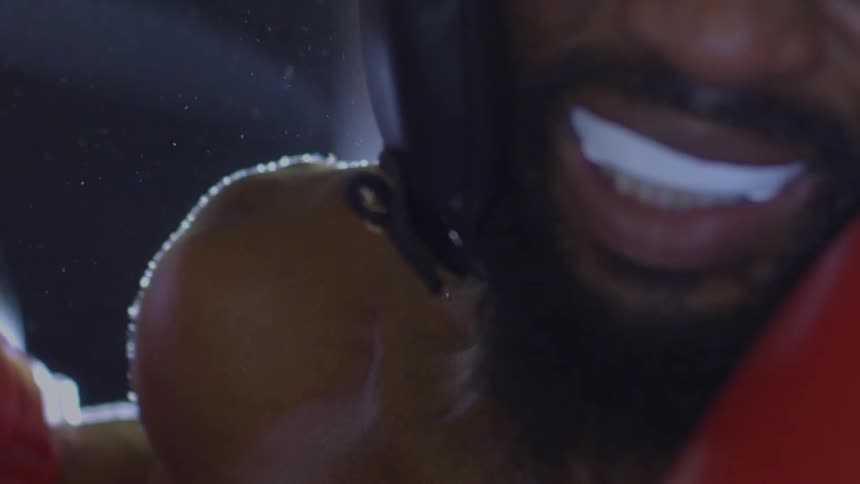 An intense, sweaty boxer wearing protective headgear and red gloves stares down the camera in a dramatic slow-motion close-up